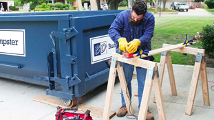 Man Cutting Wood on Driveway Next to a Dumpster