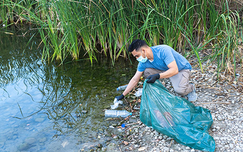 A volunteer in a mask and gloves picking up trash at a river. 